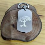 Pingente Mania de Metal com árvore gravada em metal, sobre base de madeira.