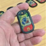 Dog tag Color com a frase "O que fazemos em vida ecoa pela eternidade", em mão sobre mesa de madeira.