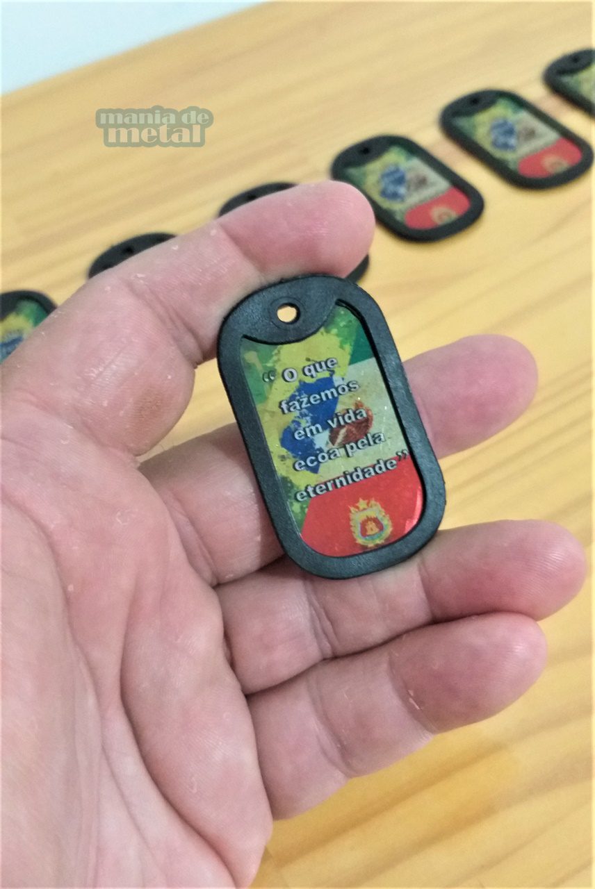 Dog tag Color com a frase "O que fazemos em vida ecoa pela eternidade", em mão sobre mesa de madeira.