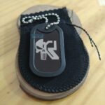 Dog tag Color preta com design de caveira e letra "K" em um suporte de madeira.
