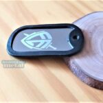 Dog Tag Fina de metal preto com design gravado, em fundo de madeira.