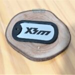 Dog tag color prata com borda preta e a inscrição "X3m".