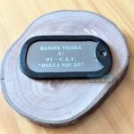 Dog Tag Fina com nome, tipo sanguíneo e esquadrão gravados sobre base de madeira rústica.