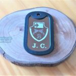 Dog Tag Fina com brasão e as letras J. C. em base de madeira rústica.