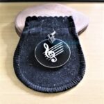 Placa Pingente Redondo em Aço Inox 316L com gravação de clave de sol e pauta musical, dentro de uma embalagem de veludo.