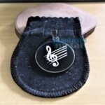 Placa Pingente Redondo: Pingente em aço inox 316L com gravação de nota musical, dentro de uma bolsa preta.
