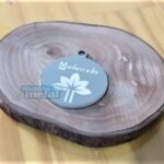 Placa Pingente Redondo em Aço Inox 316L: pingente redondo gravado com flor, sobre madeira rústica.